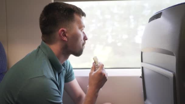 Homem viajante caucasiano viaja em um trem de alta velocidade de passageiros no assento da janela e come rolo de sanduíche de fast food. Interior do carro ferroviário. Homem viajando sozinho de trem e comendo lanche. Comida de plástico. — Vídeo de Stock