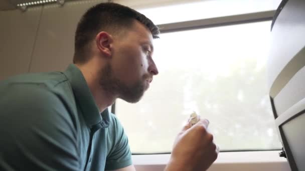 Głodny facet je fast foody w wagonie kolejowym siedzącym przy oknie. Podróż koleją. Zły nawyk. Śmieciowe żarcie. Człowiek podróżuje pociągiem i je przekąski, bułki lub kanapki. — Wideo stockowe
