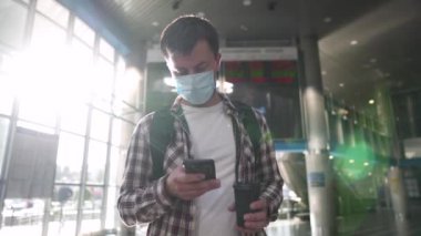 Corona covid 19 virüsü sırasında yüz maskesi takan bir adam terminalde uçuş panosunun önünde bilgi okuyor ve gitmek için telefon ve kahve kullanıyor. Ukrayna, Kiev 10 Eylül 2020