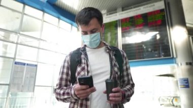 Koruyucu maskeli ve fincanlı bir adam havalimanı terminalinde, kalkış zamanı zaman çizelgesi ekran görüntüsü karantina covid 19, korona virüsü sırasında çevrimiçi check-in için akıllı telefon kullanıyor.