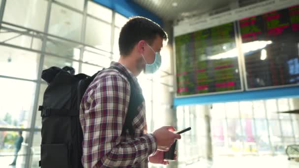 Muž v ochranné masce a šálku kávy používat chytrý telefon pro on-line check-in stojící v letištním terminálu v blízkosti jízdního řádu příletu displej obrazovky během karantény covid 19, corona virus — Stock video