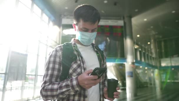 Man i skyddande medicinsk mask dricka kaffe för att gå och använda telefon på flygplatsen informationstavla på bakgrunden. Flygresekoncept under coronavirus covid 19. Förebyggande av social avståndspandemi — Stockvideo