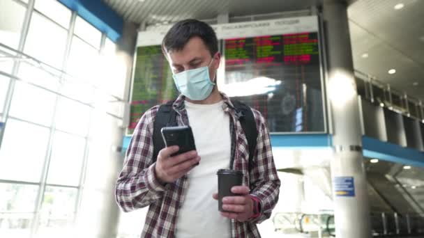 Man in beschermende medische masker drinken van koffie te gaan en het gebruik van de telefoon in de luchthaven informatiebord op de achtergrond. Vliegreis concept tijdens coronavirus covid 19. Sociale afstandspreventie pandemie — Stockvideo