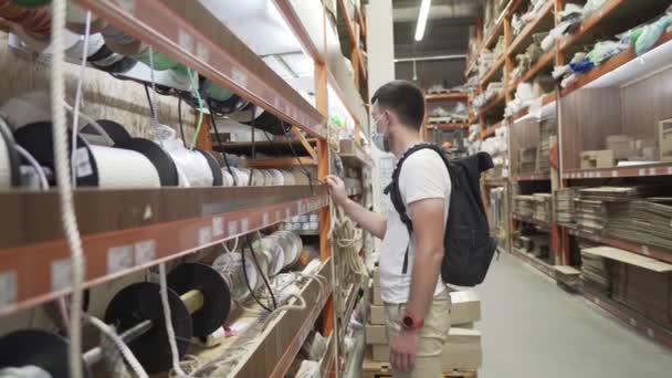 Caucasian male shopper at a hardware store, wearing a protective medical mask on his face, selects a rope from a store shelf during the corona virus and covid 19 pandemic. Building, buying — Stock Video