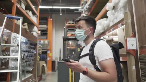 Seorang pria Kaukasia mengenakan T-shirt putih dan ransel hitam mencari pasokan di toko perangkat keras, mengenakan masker medis selama epidemi COVID-19. Topik belanja selama pandemi. — Stok Video