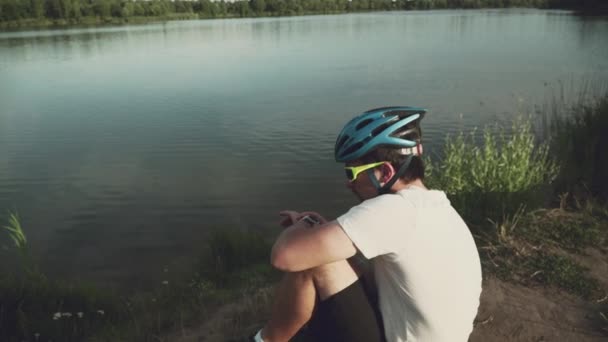 A man cyclist in a protective helmet and sunglasses sports goggles uses a wrist smartwatch after a workout while sitting in nature on the shore of a lake on a sunny day. The theme of an active — Stock Video