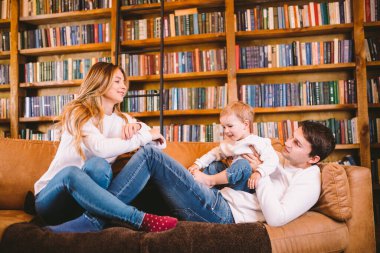 Noel aile portresi. Aynı kıyafetleri içinde genç beyaz bir aile oturma odasındaki kanepede büyük bir kitap kütüphanesi ve Noel ağacı ile eğleniyor. Mutlu yıllar ve mutlu Noeller..