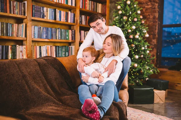 Happy parents playing with small child at Christmas time at home in living room on sofa near large cabinet with books and Christmas tree in evening. Family holiday in winter new year and christmas.