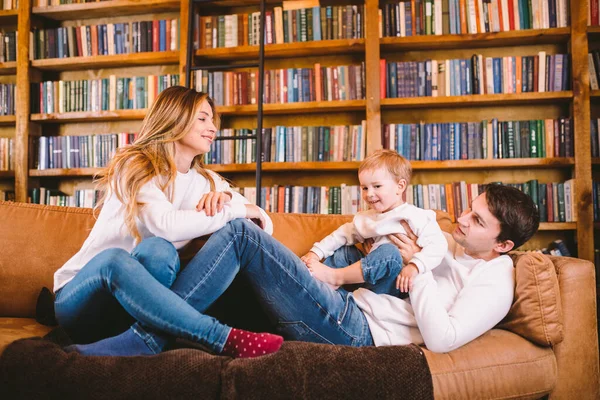 Happy parents playing with small child at Christmas time at home in living room on sofa near large cabinet with books and Christmas tree in evening. Family holiday in winter new year and christmas.