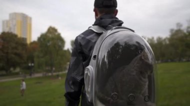 Hayvanın dikiz aynası sahibi köpüklü bir sırt çantasıyla yürüyor ve içinde gri bir kedi yavrusu taşıyor. Evcil hayvanı için lomboz taşıyan sırt çantası kullanan bir adam. Yaratıcı şeffaf evcil hayvan çantası. Taşıyıcı solunabilir kapsül