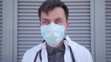 Ciddi bir doktor, tıbbi maskeyi yüzünden çıkarıyor. Hastanenin dışında dinlenen yorgun genç bir doktor. Tıp ve sağlık konsepti. Tıbbi önlüklü bir adam zor bir iş gününden sonra koruyucu maskesini çıkarıyor.