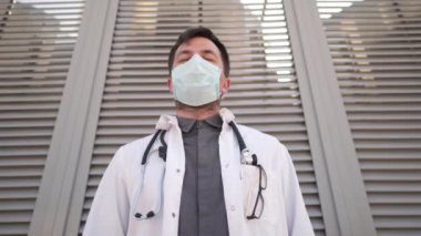 Ciddi bir doktor, tıbbi maskeyi yüzünden çıkarıyor. Hastanenin dışında dinlenen yorgun genç bir doktor. Tıp ve sağlık konsepti. Tıbbi önlüklü bir adam zor bir iş gününden sonra koruyucu maskesini çıkarıyor.