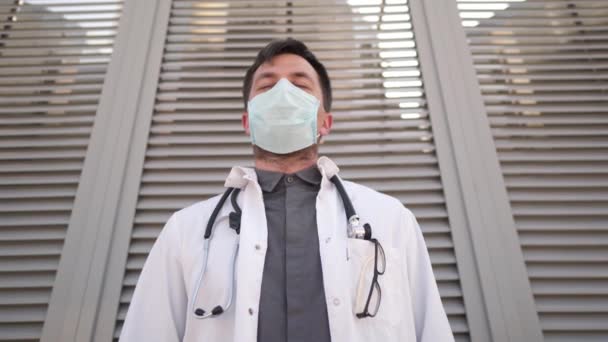 Médico serio quitando máscara médica de la cara. Un joven médico cansado descansando fuera del hospital. Concepto de medicina y salud. Hombre con uniformes médicos quitándose la máscara protectora después de un duro día en el trabajo — Vídeo de stock