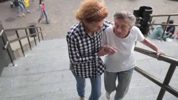 La mujer mayor y su hija de mediana edad caminan en el patio del edificio de gran altura. La enfermera ayuda a un pensionista a subir escaleras afuera. Cuidador ayudar, ayudar y apoyar a las personas mayores — Vídeo de stock