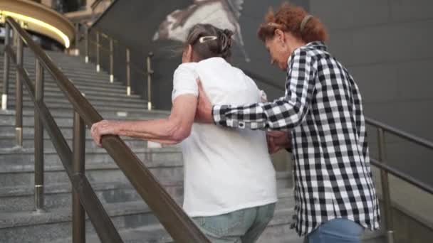 Femme âgée et sa fille d'âge moyen marchent dans la cour de l'immeuble de grande hauteur. L'infirmière aide un vieux pensionné à monter les escaliers dehors. Aide, assistance et soutien aux personnes âgées — Video