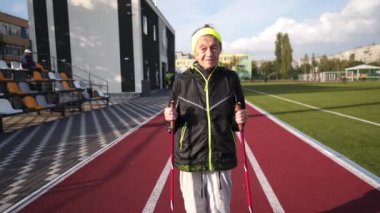 Yaşlı temasının geri kalanı aktif. Emeklilikte spor ve sağlık. Kafkas kırışıklıkları olan yaşlı bir kadın şehir stadyumunun dışında sopayla İskandinav yürüyüşü yapıyor.