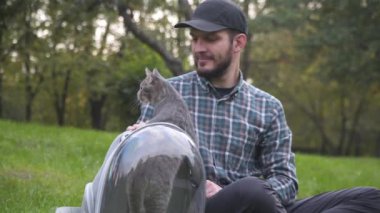 Parkta evcil hayvan taşıyıcısı olan bir adam çimenlerde oturup evcil hayvanlarla oynar. Komik kedi sırt çantasının içinde sırt çantasından dışarı bakıyor. Hayvanlarla yürümek için solunabilir kapsül.
