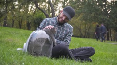 Adam, parktaki yeşil çimenlerin üzerinde oturup gri kediyle oynayıp eğlenmek için kapı koluyla sırt çantası kullanıyor. Ekipman, komik kedinin patisini sahibine doğru çektiği şeffaf nefes kapsülüdür.