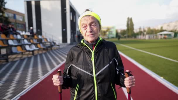 スタジアムのゴムトレッドミルでノルディックウォーキングを練習する老婦人。スカンディナヴィア人の歩行による高齢女性の歩行では、トレッキングスティックとノルディックポールを使用する。退職者の健康的なライフスタイル — ストック動画