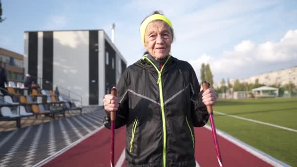 Idosas idosas caucasianas ativas de 90 anos praticam caminhada nórdica com bastões de esqui em uma pista com revestimento de borracha vermelha. Férias activas. Uma mulher em forma. Estilo de vida saudável para os idosos — Vídeo de Stock