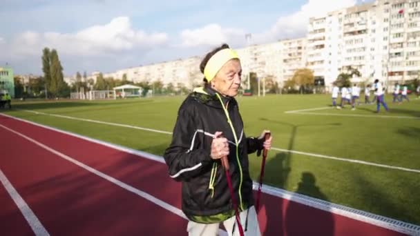 Descanso activo del tema de los ancianos. Deportes y salud en la jubilación. Mujer caucásica muy anciana con arrugas profundas haciendo ejercicios de caminata nórdica con palos afuera en el estadio de la ciudad — Vídeos de Stock