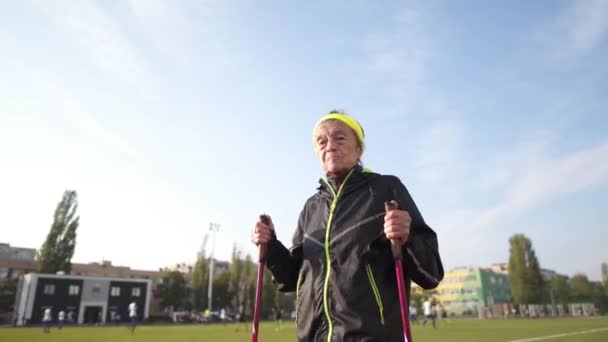 Mujer mayor caminando con bastones en el estadio en una cubierta de goma roja. Mujer mayor de 88 años haciendo ejercicios de senderismo nórdico en el estadio de la ciudad en la pista de atletismo. concepto de estilo de vida saludable — Vídeos de Stock