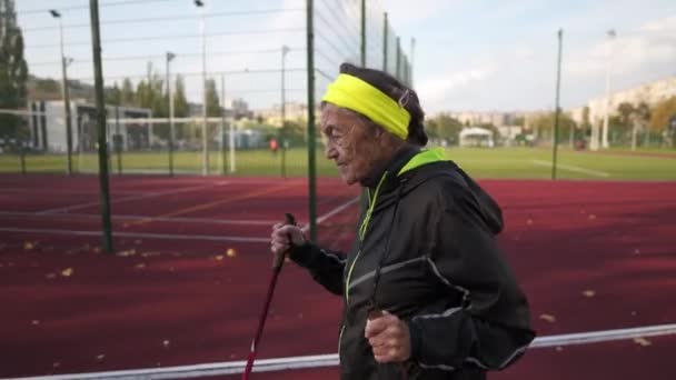 Egy öregasszony, aki a skandináv sétát gyakorolja a stadionban a gumi futópadon. Az idősebb női séta a skandináv séta használja túrabotok és északi pólusok. Nyugdíjasok egészséges életmódja — Stock videók