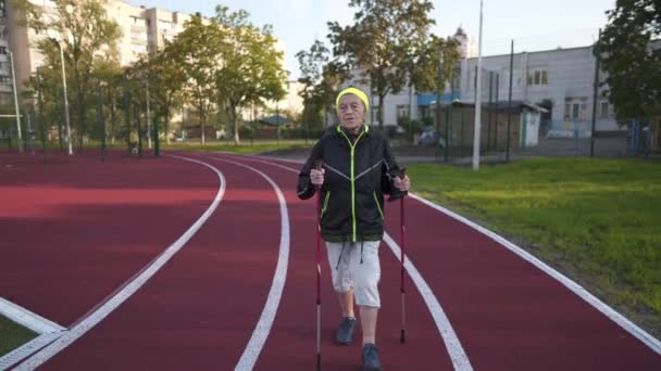 Resto ativo do tema dos idosos. Esportes e saúde na aposentadoria. Mulher muito velha caucasiana com rugas profundas fazendo exercícios de caminhada nórdica com paus fora no estádio da cidade — Vídeo de Stock