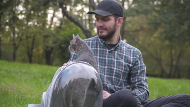 Férfi díszes kisállat hordozó szabadban parkban ül a gyepen, és játszik kisállat. Vicces macska néz ki hátizsák -ban átlátszó kisállat hátizsák. Hordozó belélegezhető kapszula az állatokkal való gyalogláshoz — Stock videók