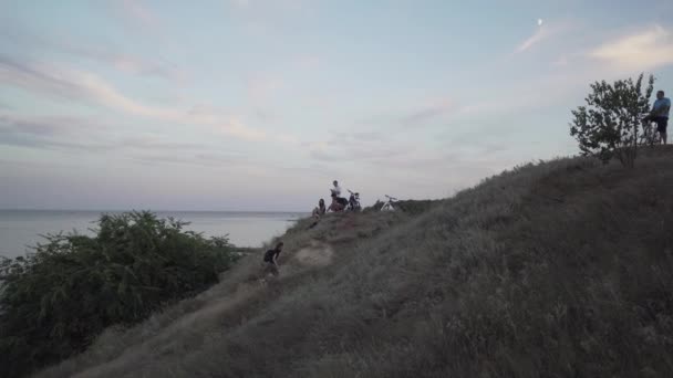 Украина, Одесса. 12 августа 2020.Группа кавказских молодых людей с велосипедами отдыхают на вершине холма у берега моря теплым летним вечером, мужчина бежит по холму, вид на горизонт. Тема морского отдыха. — стоковое видео