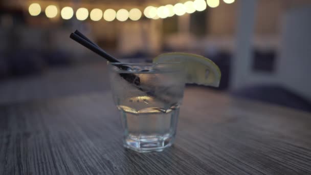 En kall alkoholhaltig dryck av transparent färg i ett glas med lågt glas med citronskiva och två sugrör på ett träbord i en restaurang runt en mörk och retrobelysning, barkonst. — Stockvideo