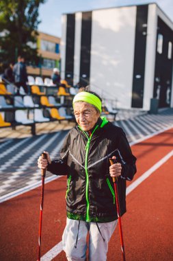 Yaşlı temasının geri kalanı aktif. Emeklilikte spor ve sağlık. Kafkas kırışıklıkları olan yaşlı bir kadın şehir stadyumunun dışında sopayla İskandinav yürüyüşü yapıyor..