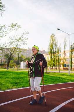 Yaşlı temasının geri kalanı aktif. Emeklilikte spor ve sağlık. Kafkas kırışıklıkları olan yaşlı bir kadın şehir stadyumunun dışında sopayla İskandinav yürüyüşü yapıyor..