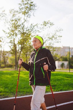 90 yıllık Aktif Kafkasyalı yaşlı kadınlar, kırmızı kauçuk kaplamalı bir pistte kayak direkleriyle İskandinav yürüyüşü yapıyorlar. Aktif tatiller. Formda bir kadın. Yaşlılar için sağlıklı bir yaşam tarzı.