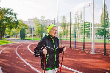 Yaşlı temasının geri kalanı aktif. Emeklilikte spor ve sağlık. Kafkas kırışıklıkları olan yaşlı bir kadın şehir stadyumunun dışında sopayla İskandinav yürüyüşü yapıyor..