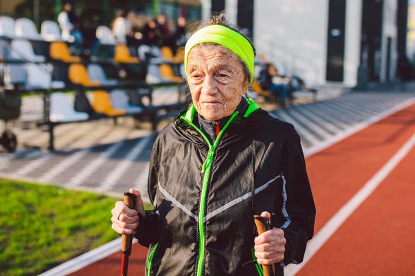 Active elderly Caucasian elderly women of 90 years practice Nordic walking with ski poles on a track with a red rubber coating. Active holidays. A fit woman. Healthy lifestyle for the elderly.