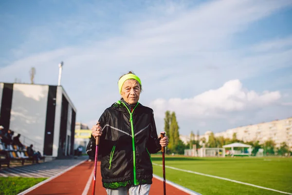 Senior woman walking with walking poles in stadium on a red rubber cover. Elderly woman 88 years old doing Nordic walking exercises at the city stadium on the running track. Healthy lifestyle concept.