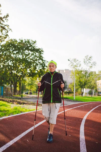 Senior woman walking with walking poles in stadium on a red rubber cover. Elderly woman 88 years old doing Nordic walking exercises at the city stadium on the running track. Healthy lifestyle concept.