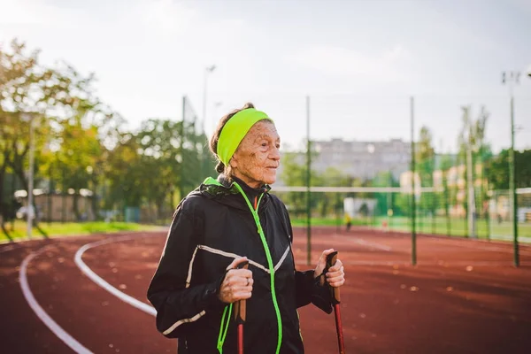 Senior woman walking with walking poles in stadium on a red rubber cover. Elderly woman 88 years old doing Nordic walking exercises at the city stadium on the running track. Healthy lifestyle concept.