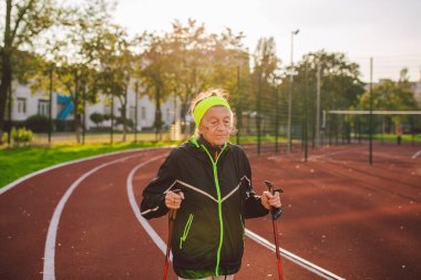 Yaşlı temasının geri kalanı aktif. Emeklilikte spor ve sağlık. Kafkas kırışıklıkları olan yaşlı bir kadın şehir stadyumunun dışında sopayla İskandinav yürüyüşü yapıyor..