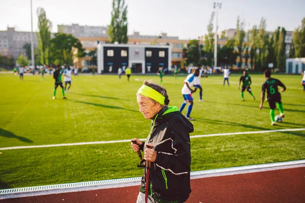 Active rest of the elderly theme. Sports and health in retirement. Caucasian very old woman with deep wrinkles doing Nordic walking exercises with sticks outside in the city stadium.