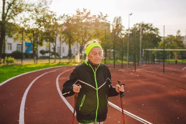 Active rest of the elderly theme. Sports and health in retirement. Caucasian very old woman with deep wrinkles doing Nordic walking exercises with sticks outside in the city stadium.