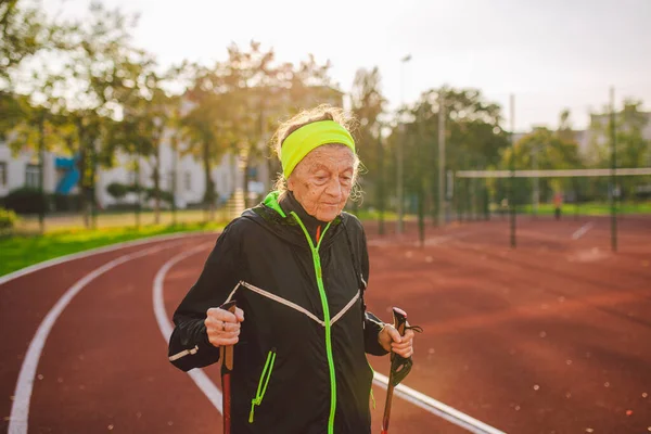 Senior woman walking with walking poles in stadium on a red rubber cover. Elderly woman 88 years old doing Nordic walking exercises at the city stadium on the running track. Healthy lifestyle concept.