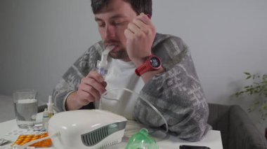 Genç bir adam, boğaz ağrısı ve öksürükten muzdarip, battaniyeye sarılı olarak evinde oturuyor. Nebulizör kullanıyor. Soğuk algınlığı, bronşit ve astım belirtilerini tedavi etmek için tıbbi bir cihaz.