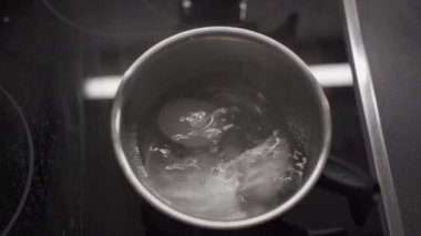 İki beyaz tavuk yumurtası ev mutfağındaki elektrikli ocakta küçük metalik bir tencerede kaynıyor. Yemek pişirme, kahvaltı hazırlama, protein yiyeceği, sağlıklı beslenme, beslenme ve aşçılık süreci kavramı.