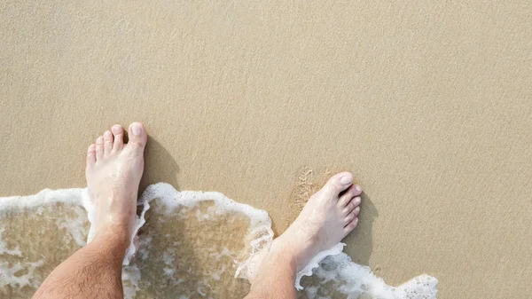 Closeup of a man\'s bare feet stand at wet on the beach , with a wave\'s edge foaming gently beneath them. Vacation on ocean beach, foot on sea sand. Leave empty copy space Enter the text above.