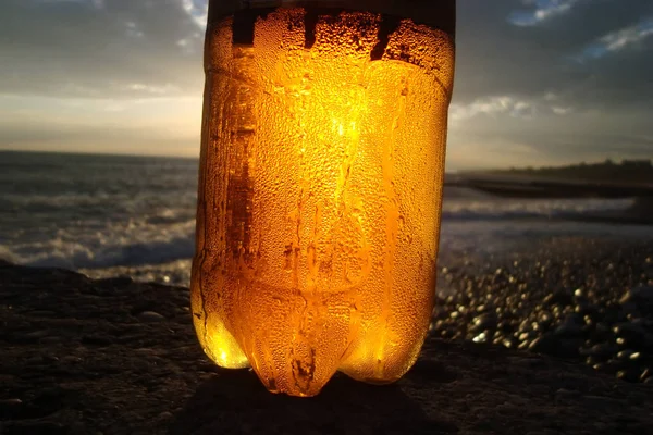 Plastic beer bottle with drops of water, against the sunset of the sea.