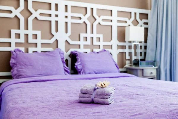 Small violet towels decorated with the flower laying on the bed with pillows and violet cover in cozy bedroom interior