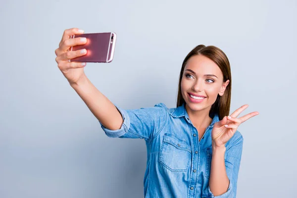 Portrait of trendy stylish woman shooting self portrait on smart phone gesturing v-sign wearing jeans shirt isolated on grey background