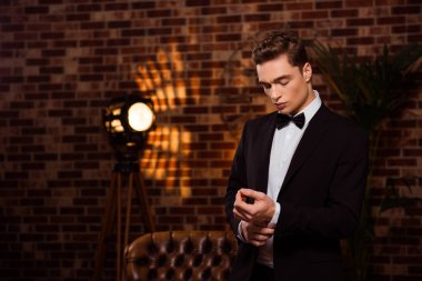 Horizontal portrait of manly, harsh, cool, virile, stunning, perfect man in black suit with bow, preparing for date, buttoning cuffs on sleeve of white shirt over brick wall clipart
